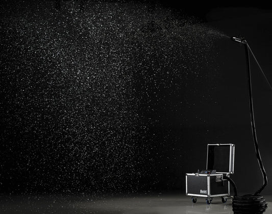 The Antari S-500 900 W Snow Machine by Antari glides on wheels, spraying a fine mist of snow across a dark, empty space, the droplets beautifully illuminated against the black backdrop.