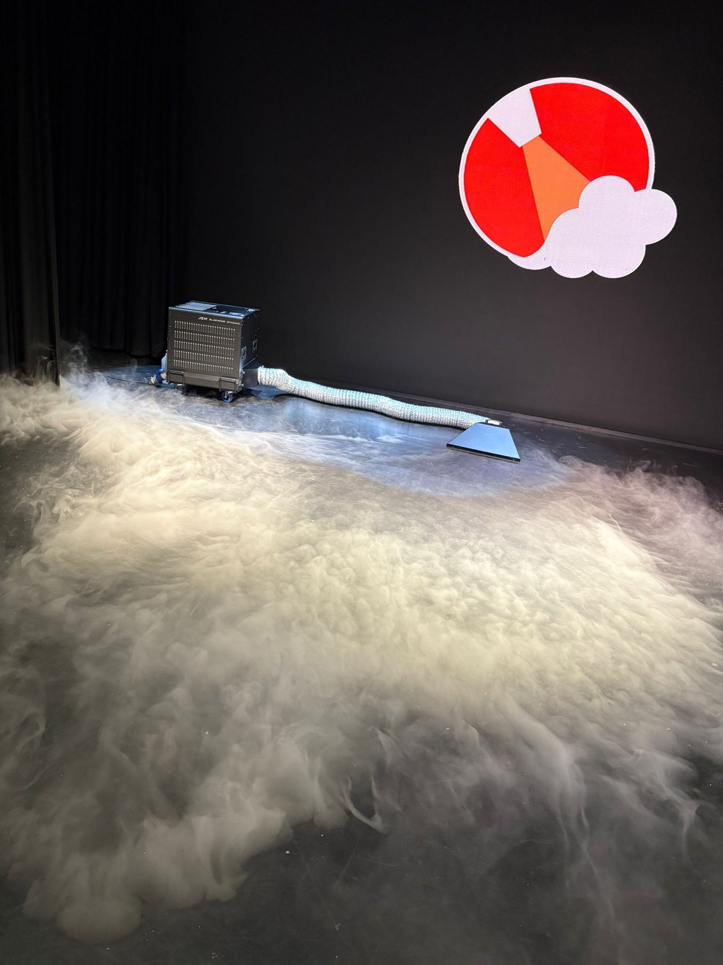 The JEM Martin Glaciator Dynamic Fog Blade generates dense, low-lying fog across a dark room’s floor, while the stage’s back wall features a vivid graphic of a red and orange rocket launching over white clouds.