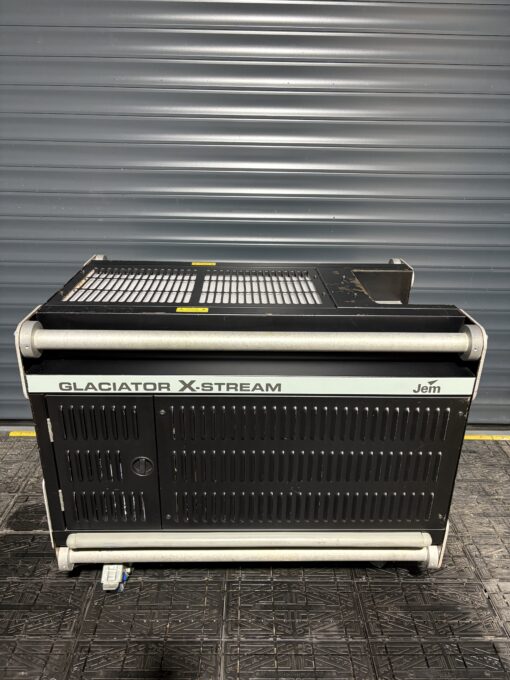 A large, black JEM Professional Glaciator X-Stream low-fog generator from JEM Professional sits on a textured floor in front of a gray metal roller shutter door, ready to create impressive ground-hugging and heavy-fog effects.