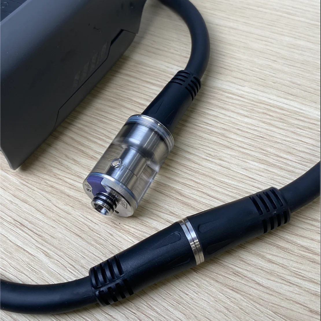 A close-up of the PMI SmokeNINJA Extension Cable, featuring black connectors, rests on a light wood surface next to a dark rectangular device, likely part of a fog chamber or smoke delivery system.