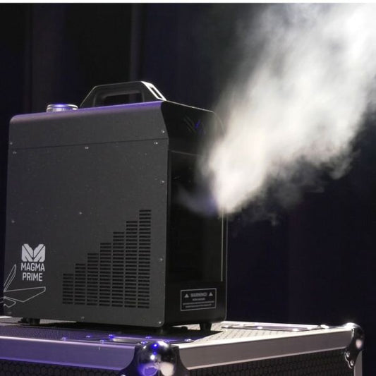 The Elation-Magmatic Magma Prime Hazer 700W emits a thick stream of haze, shown resting on a hard case against a dark background.