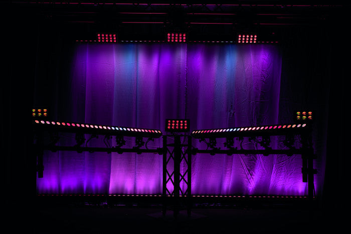 A stage lighting setup with purple and pink illumination highlights a curtain backdrop, featuring an array of Showtec Cameleon Bar 15 Q6 Tour RGBWA-UV LED Bars and beams arranged on stands in a dark environment.
