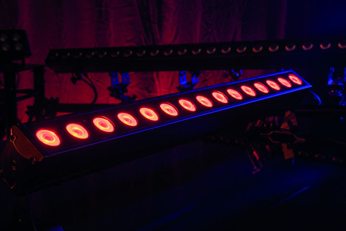A row of vibrant red stage lights from the Showtec Cameleon Bar 15 Q6 Tour RGBWA-UV LED Bar illuminates a dark room with a purple backdrop, courtesy of Showtec, creating a dramatic and moody atmosphere.