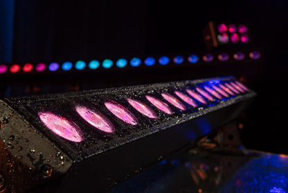 A row of illuminated Showtec Cameleon Bar 15 Q6 Tour RGBWA-UV LED Bars glows with vibrant pink and purple hues, water droplets beading on the surface to emphasize the IP65-rated durability. In the background, another softly focused row of blue and pink Showtec RGBWA-UV LED Bars showcases the vivid, versatile effects possible with this fixture.