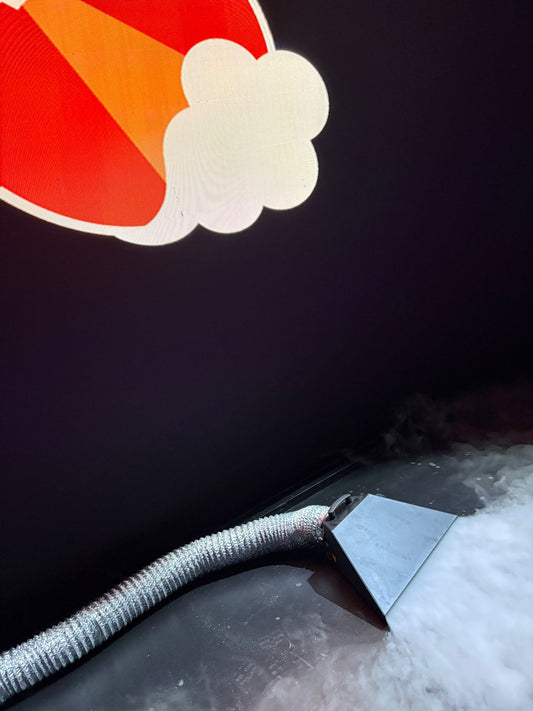 A close-up of a vacuum cleaner hose with a wide nozzle sucking up white foam from a dark surface; above, a bright cartoon image is projected, suggesting low-fog output similar to the JEM Martin Glaciator Dynamic Fog Blade.