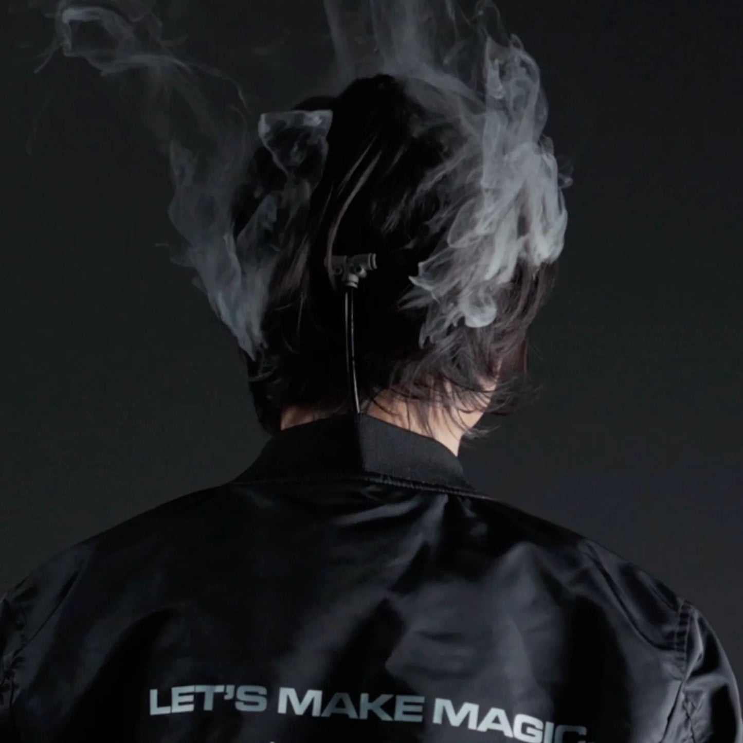 A person with dark hair, seen from behind, wears the PMI SmokeNINJA PRO/NINJA/GENIE Smoke Vest (PMI_AC_SVEST) by PMI as swirling smoke surrounds them. They sport a black jacket reading "LETS MAKE MAGIC" on a dark background.