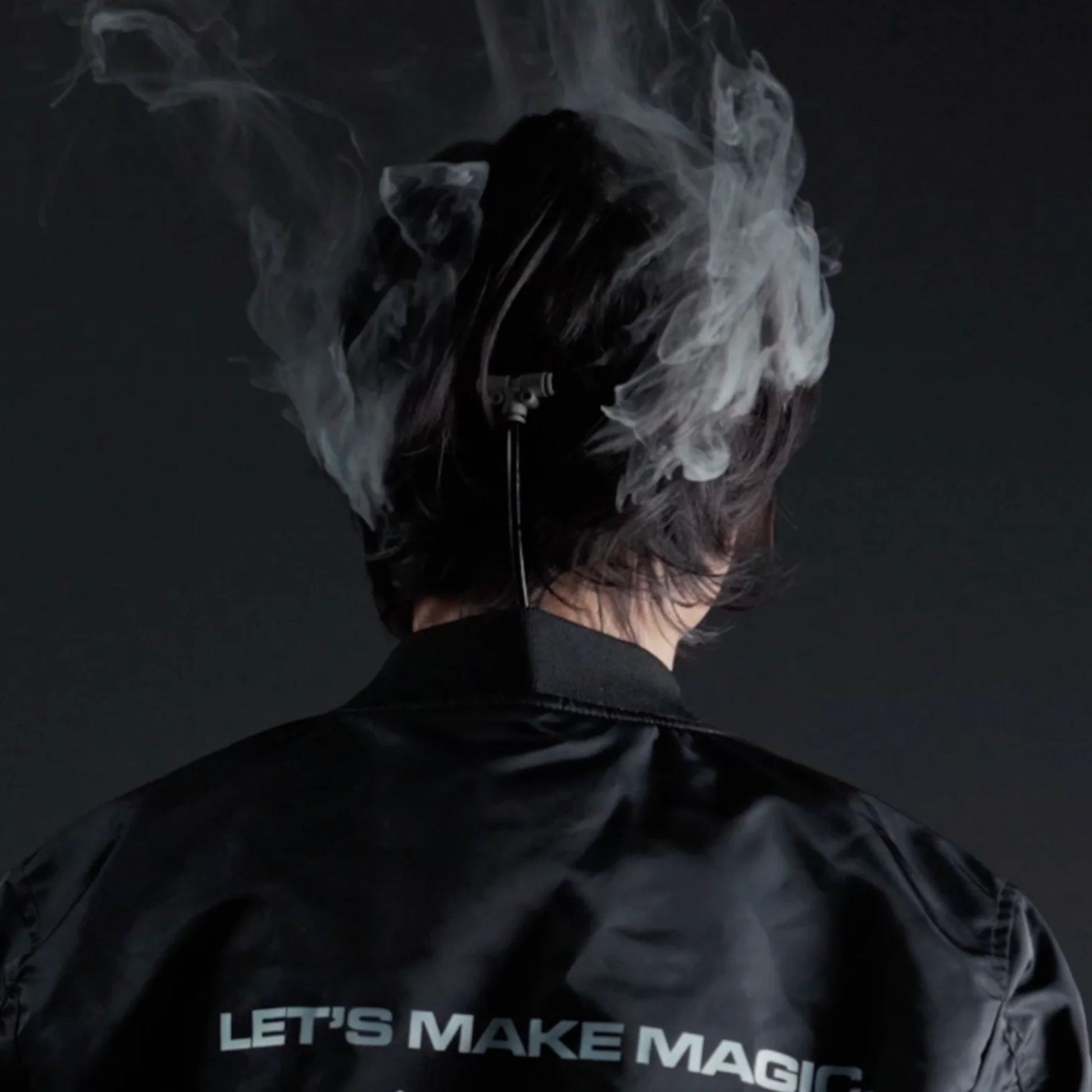 A person with dark hair, seen from behind, wears the PMI SmokeNINJA PRO/NINJA/GENIE Smoke Vest (PMI_AC_SVEST) by PMI as swirling smoke surrounds them. They sport a black jacket reading "LETS MAKE MAGIC" on a dark background.