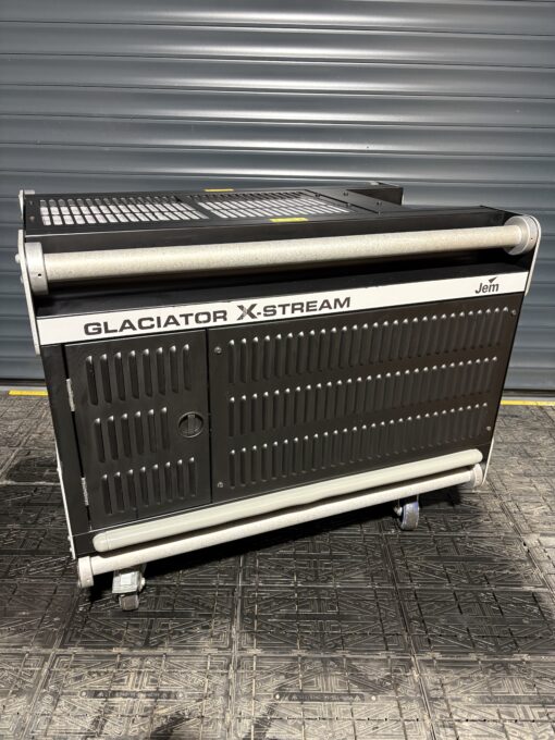 A JEM Professional Glaciator X-Stream low-fog generator machine by JEM Professional, finished in black and mounted on wheels, stands on a textured floor in front of a gray metal roller door, ready to create dramatic ground-hugging fog effects.