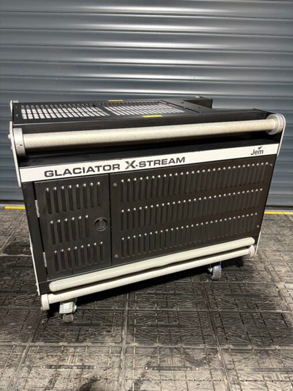A JEM Professional Glaciator X-Stream low-fog generator machine by JEM Professional, finished in black and mounted on wheels, stands on a textured floor in front of a gray metal roller door, ready to create dramatic ground-hugging fog effects.