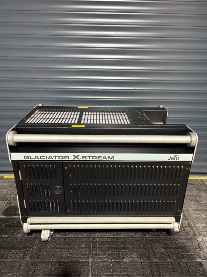 A large, black JEM Professional Glaciator X-Stream low-fog generator from JEM Professional sits on a textured floor in front of a gray metal roller shutter door, ready to create impressive ground-hugging and heavy-fog effects.
