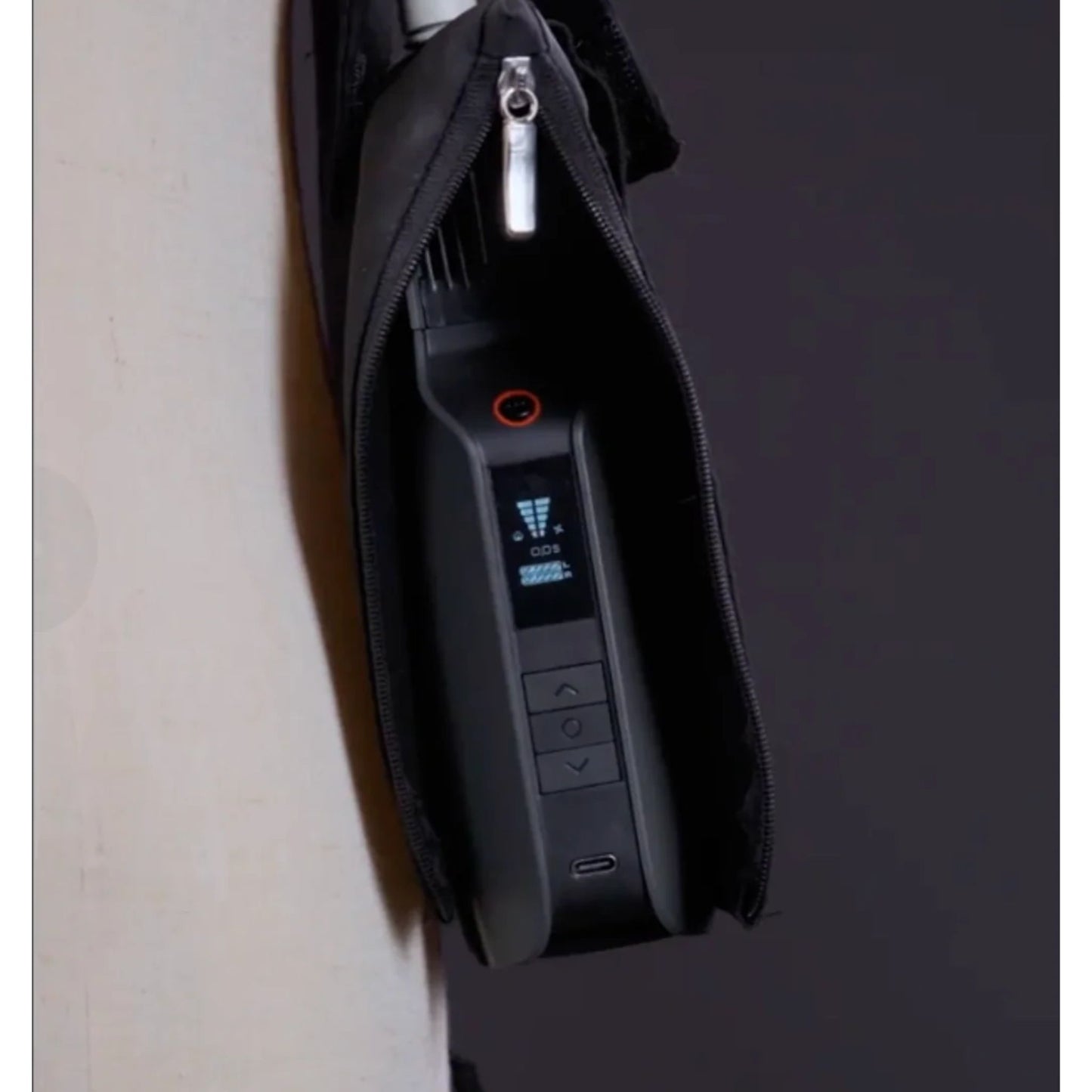A close-up of the PMI SmokeNINJA PRO/NINJA/GENIE PMI Smoke Vest (PMI_AC_SVEST), featuring a black electronic device with a small screen and buttons, partially zipped inside its black case, hanging from a white surface.