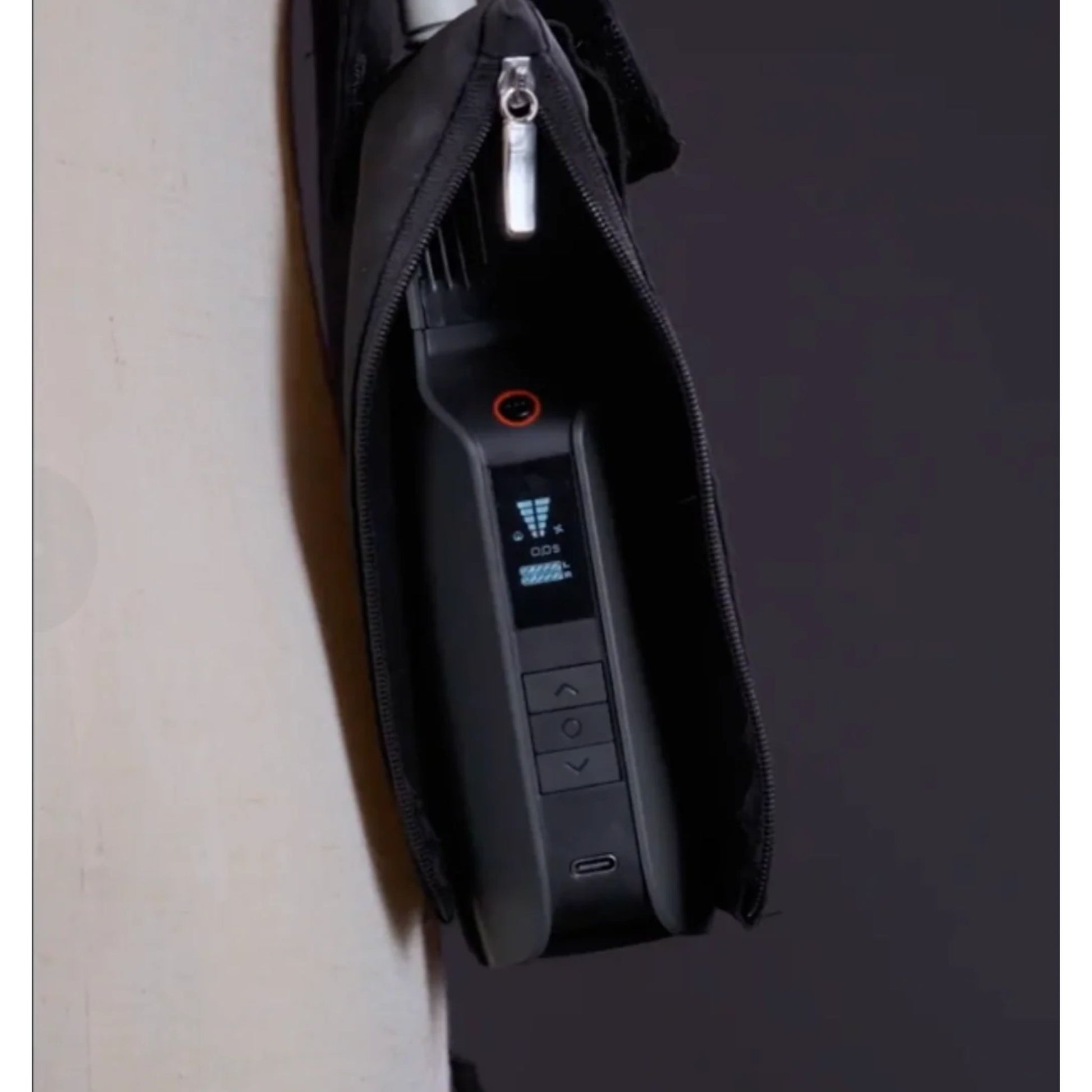 A close-up of the PMI SmokeNINJA PRO/NINJA/GENIE PMI Smoke Vest (PMI_AC_SVEST), featuring a black electronic device with a small screen and buttons, partially zipped inside its black case, hanging from a white surface.