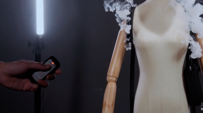 A person holds a remote near a mannequin with smoke rising from its wooden shoulder, demonstrating the PMI SmokeNINJA PRO/NINJA/GENIE PMI Smoke Vest against a dark background with bright light on the left.