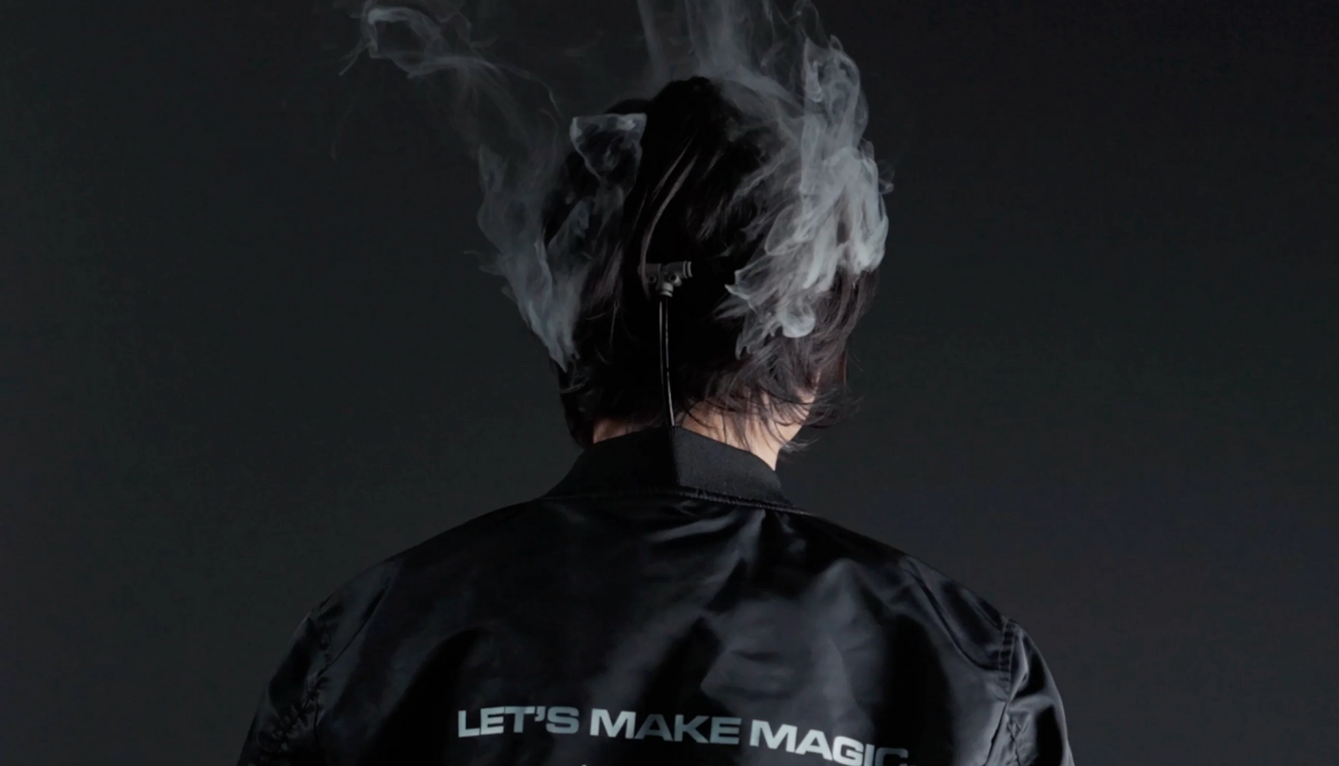 Seen from behind, a person with dark hair wears a black jacket reading "LET'S MAKE MAGIC" while smoke from the PMI SmokeNINJA PRO/NINJA/GENIE PMI Smoke Vest rises above their head against a dark background.