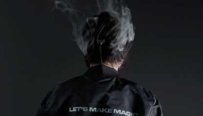 Seen from behind, a person with dark hair wears a black jacket reading "LET'S MAKE MAGIC" while smoke from the PMI SmokeNINJA PRO/NINJA/GENIE PMI Smoke Vest rises above their head against a dark background.