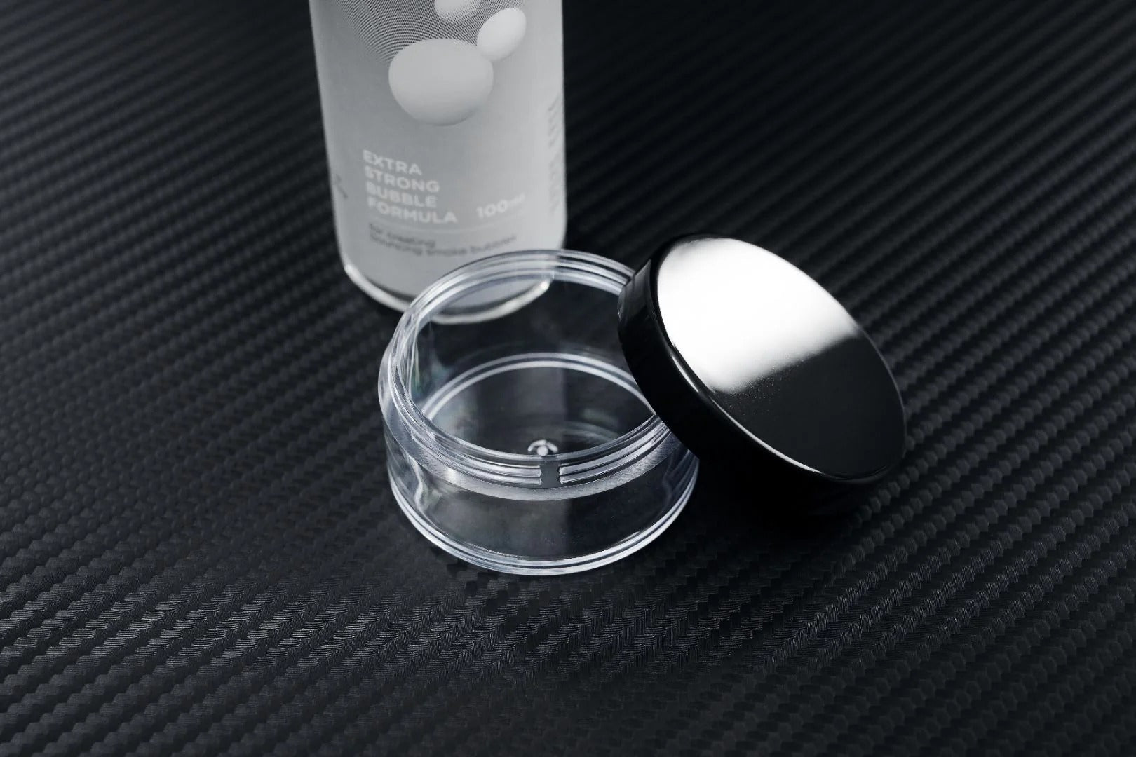 A clear, empty cosmetic jar with a black lid rests open on textured black next to a partially visible PMI SmokeNINJA Bubble Kit 100ml bottle labeled EXTRA STRONG FORMULA—evoking memories of wedding moments beneath snowy bubble rain.