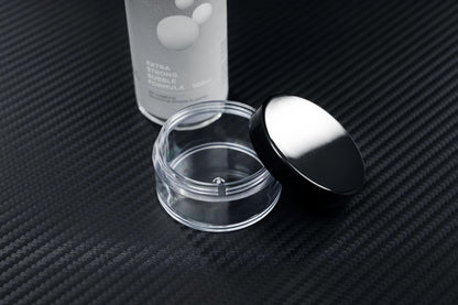 A clear, empty cosmetic jar with a black lid rests open on textured black next to a partially visible PMI SmokeNINJA Bubble Kit 100ml bottle labeled EXTRA STRONG FORMULA—evoking memories of wedding moments beneath snowy bubble rain.