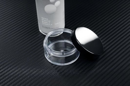 A clear, empty cosmetic jar with a black lid rests open on textured black next to a partially visible PMI SmokeNINJA Bubble Kit 100ml bottle labeled EXTRA STRONG FORMULA—evoking memories of wedding moments beneath snowy bubble rain.