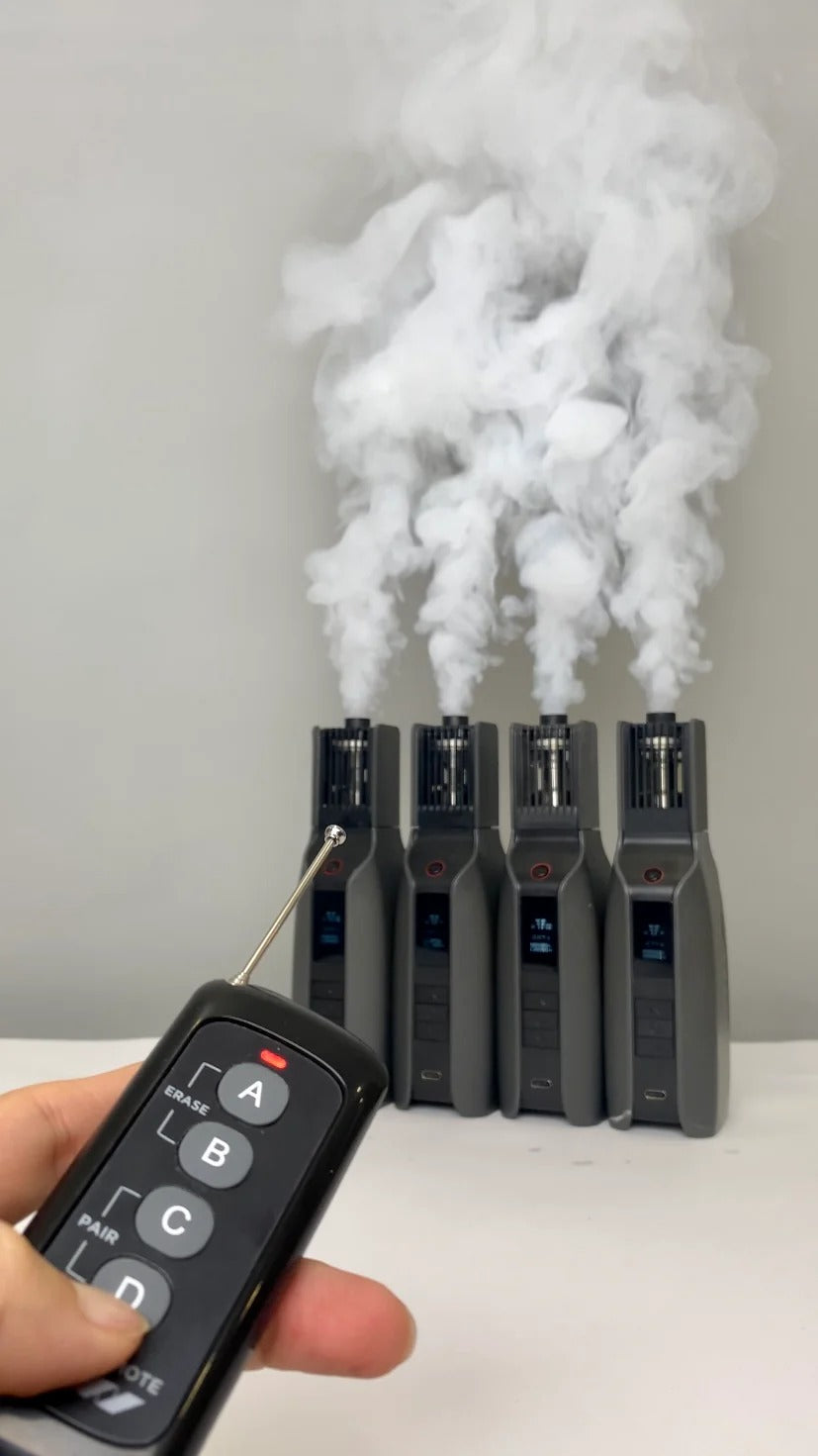 A hand holds the PMI SmokeNINJA Magic Remote labeled A, B, C, and D as four Smoke Genie machines emit thick white smoke in a row against a plain background.