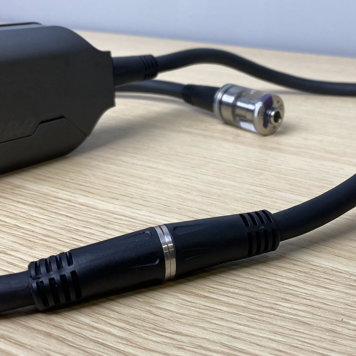 A close-up of PMI's SmokeNINJA Extension Cable, featuring black cables with metal connectors and ribbed grips, resting on a light wood surface as they connect to a partially visible black device on the left.