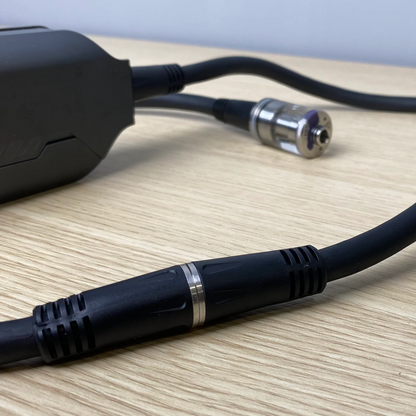 A close-up of PMI's SmokeNINJA Extension Cable, featuring black cables with metal connectors and ribbed grips, resting on a light wood surface as they connect to a partially visible black device on the left.