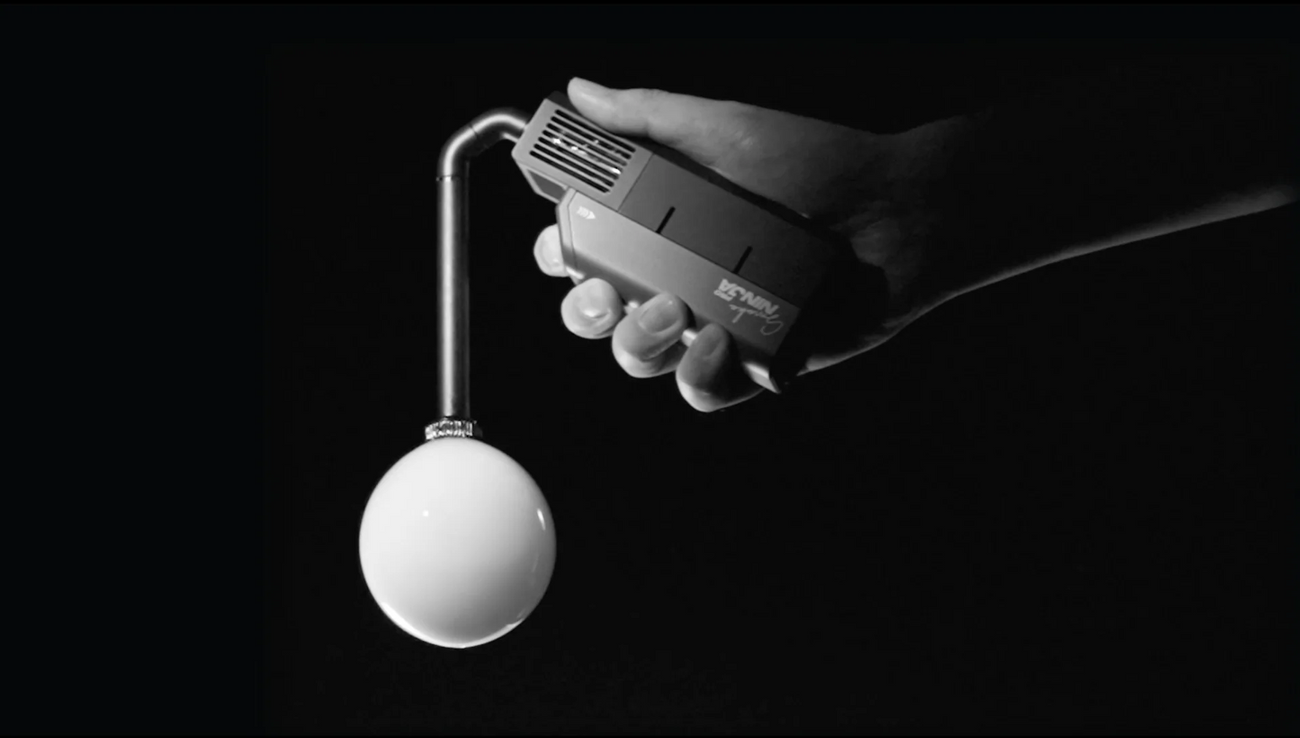 A hand holds the PMI SmokeNINJA Bubble Kit 100ml, featuring a bent metal tube with a round white bulb nozzle, set against a black background. The monochrome image evokes the playful feel of bubble-like smoke suspended in air.