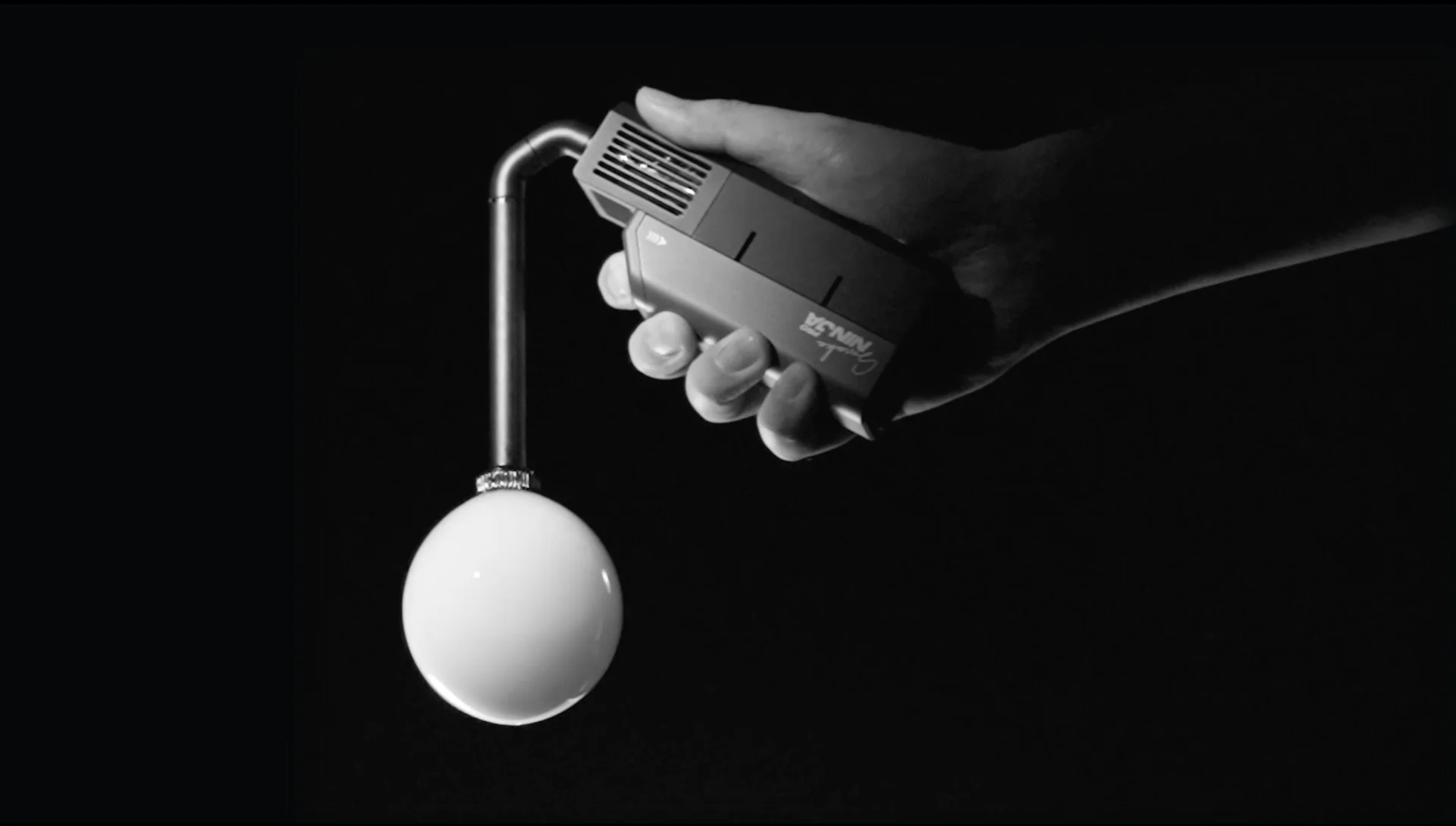 A hand holds the PMI SmokeNINJA Bubble Kit 100ml, featuring a bent metal tube with a round white bulb nozzle, set against a black background. The monochrome image evokes the playful feel of bubble-like smoke suspended in air.