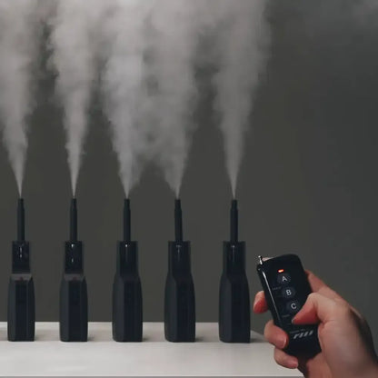 A hand uses the PMI SmokeNINJA Magic Remote to activate five Smoke Genie devices on a table, each emitting a vertical stream of white mist for seamless multi-machine control.