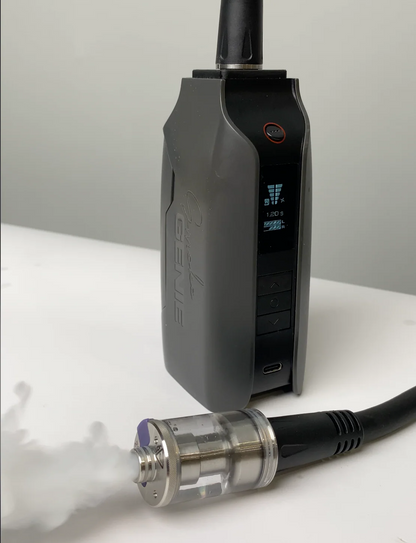 A black PMI SmokeNINJA Extension Cable is displayed upright on a white surface with its settings visible. In front, a vape tank connects via the black cable to a fog chamber emitting visible vapor.