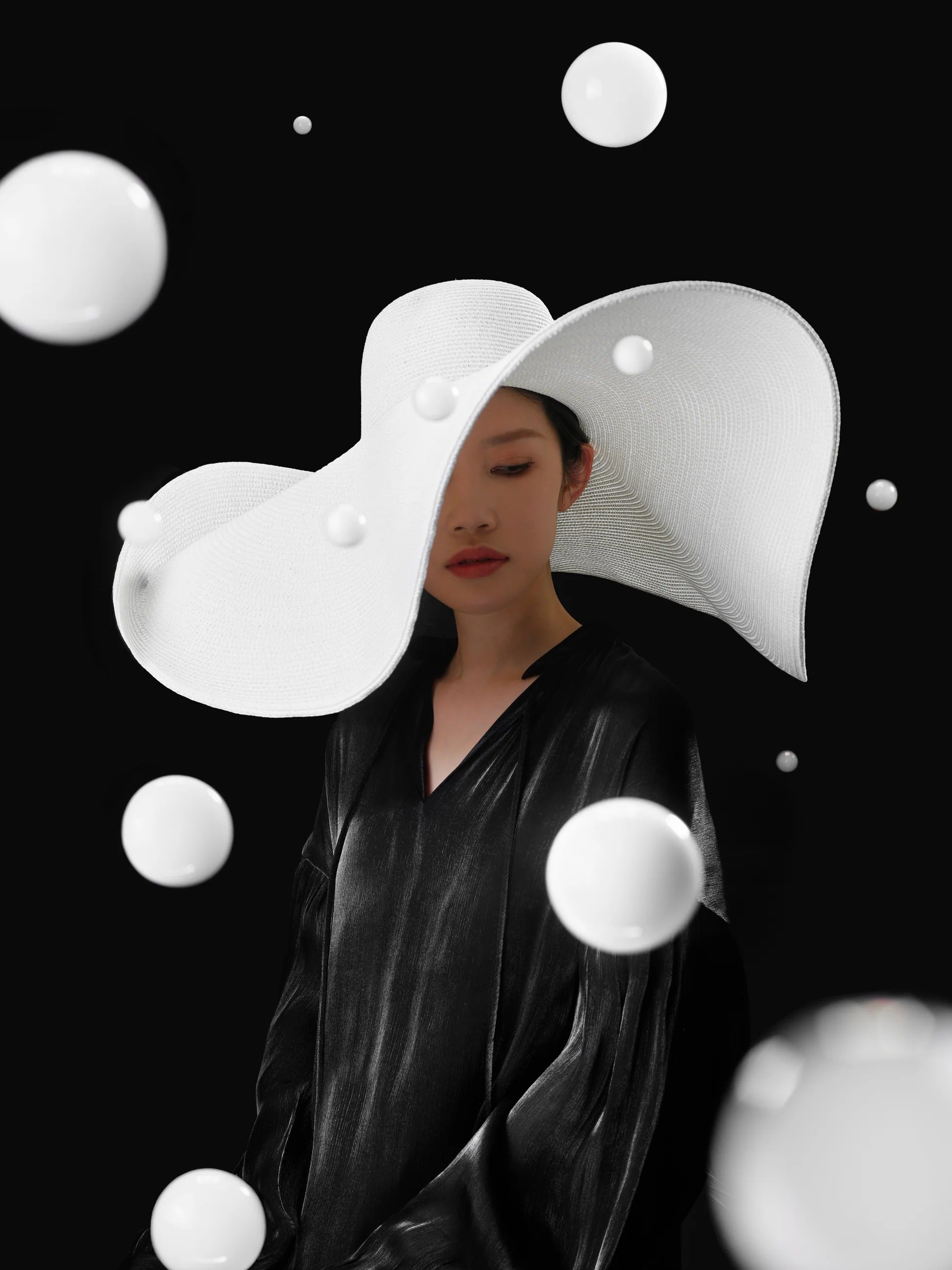 A woman in black with a sculptural white hat stands amid floating white bubbles, created by the SmokeNINJA Bubble Kit 100ml from PMI, set against a black background. Her hat casts striking shadows across her face.