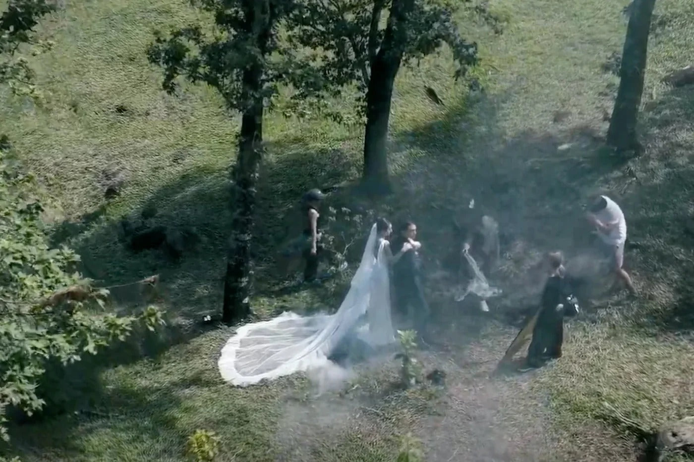 A bride in a long white veil walks on grass with attendants as trees cast shadows nearby. The dreamy, cinematic haze is created by PMI’s SmokeNINJA Pro Hazer Kit—perfect for photography and video production.